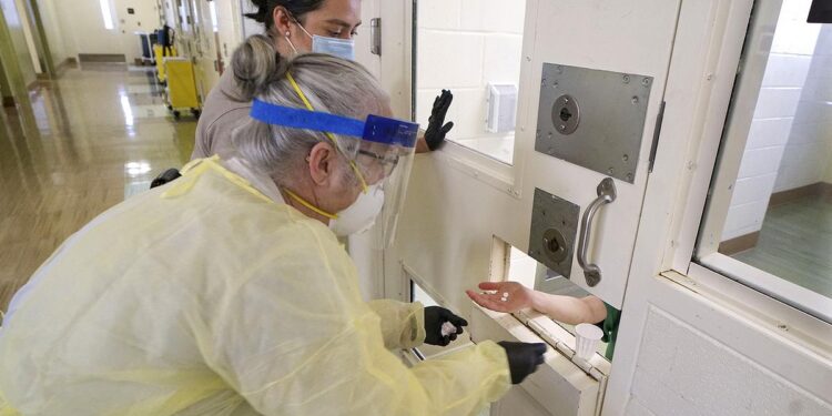 A sheriff’s deputy and a nurse stand in front of a prison door to give medication to an incarcerated person.