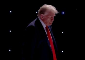 Former President Donald Trump, a White man wearing a black suit and red tie, looks down. There are small studio lights in the background.