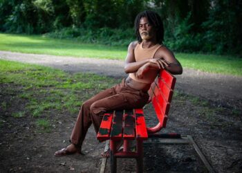 A photo of a Black person with black hair and a blond beard sitting on a red park bench while looking at the camera.