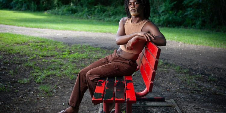 A photo of a Black person with black hair and a blond beard sitting on a red park bench while looking at the camera.