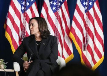 Vice President Kamala Harris, an Indian and Black woman with medium skin tone wearing a black suit, looks to the left while holding a microphone. She is sitting on a chair in front of three American flags.