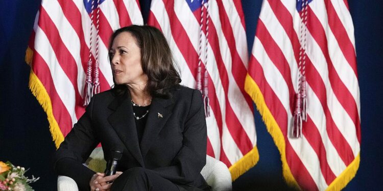Vice President Kamala Harris, an Indian and Black woman with medium skin tone wearing a black suit, looks to the left while holding a microphone. She is sitting on a chair in front of three American flags.