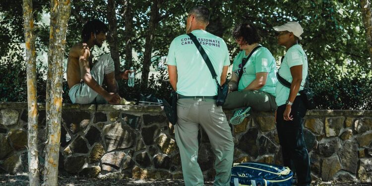 A crisis response team of three people, wearing cargo pants, turquoise shirts and fanny packs, talk to a medium-dark-skinned man sitting on a concrete wall. The man, wearing gray sweatpants, holds a water bottle in one hand.