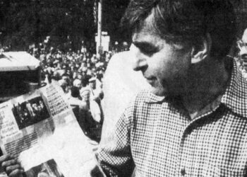 Gov. Michael Dukakis, a White man in a checkered shirt, holds a leaflet that has the words "Vote for George Bush" and "Security" printed on them, among other text that is not visible.