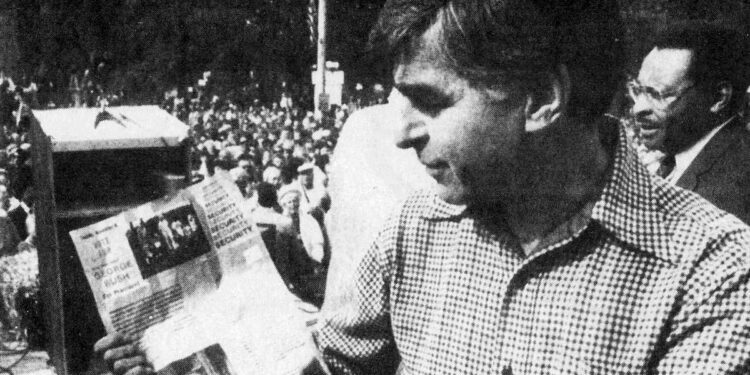 Gov. Michael Dukakis, a White man in a checkered shirt, holds a leaflet that has the words "Vote for George Bush" and "Security" printed on them, among other text that is not visible.