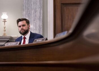 A middle-aged White man with brown hair and a beard, wearing a blue suit with a red tie, sits in front of a microphone at the end of a curved, wooden table.