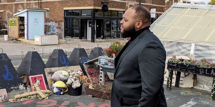 A Black man, wearing a black suit, looks to the left while standing in front of a memorial on a street.
