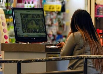 A woman with long dark hair with a gradient color and wearing a light gray long-sleeved shirt looks at security camera footage at a desk inside a store.