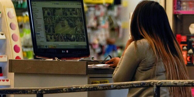 A woman with long dark hair with a gradient color and wearing a light gray long-sleeved shirt looks at security camera footage at a desk inside a store.