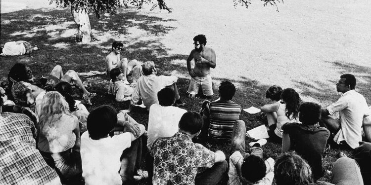 A black-and-white photo shows a bearded White man kneeling and talking to a group of about 20 people, all sitting on the grass in a park.