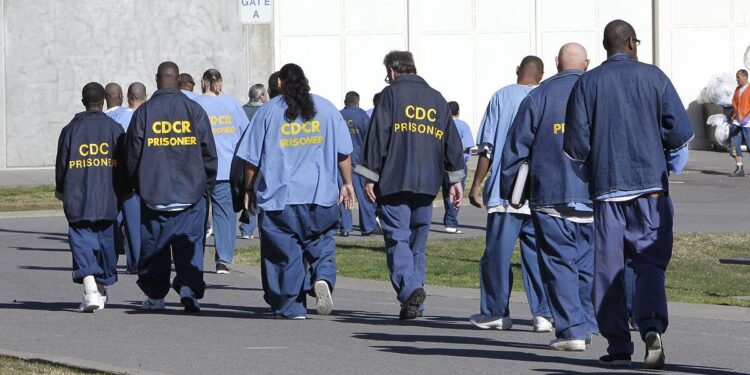 A group of people, wearing blue prison uniforms with the words "CDCR prisoner" on their backs, walk through a prison yard.