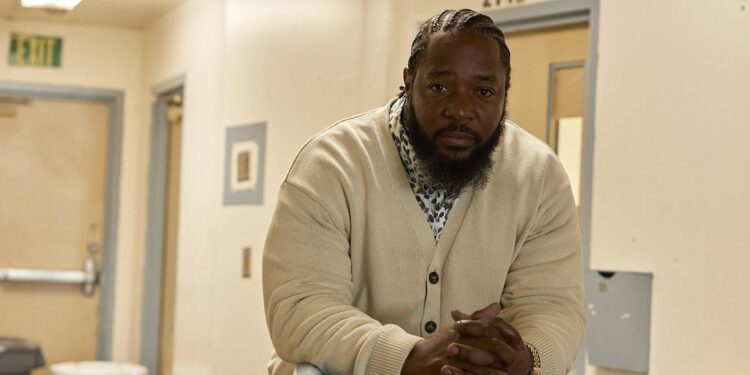 Ofonzo Staton, a Black man with a beard and goatee, wears a cream-colored cardigan and black pants. He leans against a railing painted light blue with a visitor's badge marked "11", looking towards the viewer with a pensive expression.
