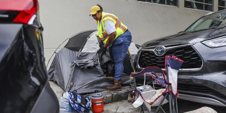 A dark-skinned woman, wearing a reflective safety vest, white T-shirt, blue jeans, yellow cap and gloves, takes items out of a tent located on a sidewalk. A camping chair, a backpack, a plastic bag and a water jug are in front of the tent. Two cars are parked next to the tent.
