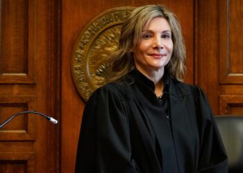 A White, blonde woman stands in a black robe behind a judge’s podium.