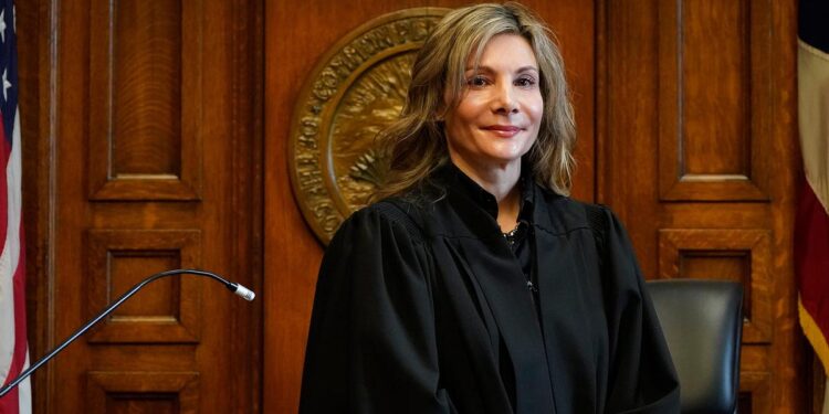 A White, blonde woman stands in a black robe behind a judge’s podium.
