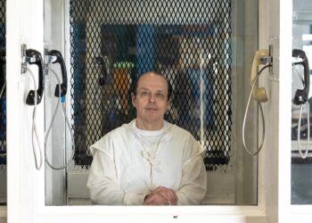 Robert Roberson, a light-skinned man wearing a white prison uniform, looks through visitation room glass. A phone hangs on either side of the divider.