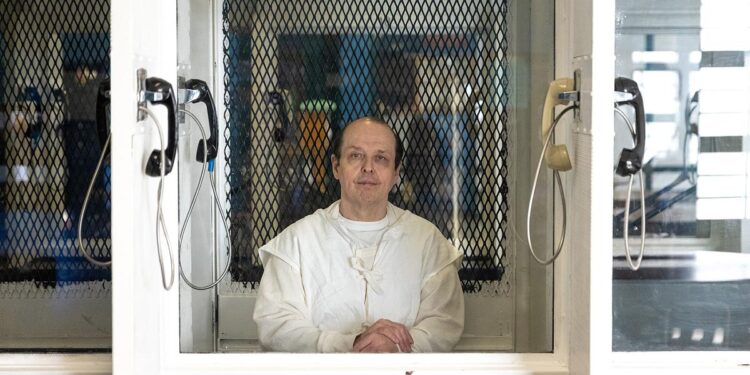 Robert Roberson, a light-skinned man wearing a white prison uniform, looks through visitation room glass. A phone hangs on either side of the divider.