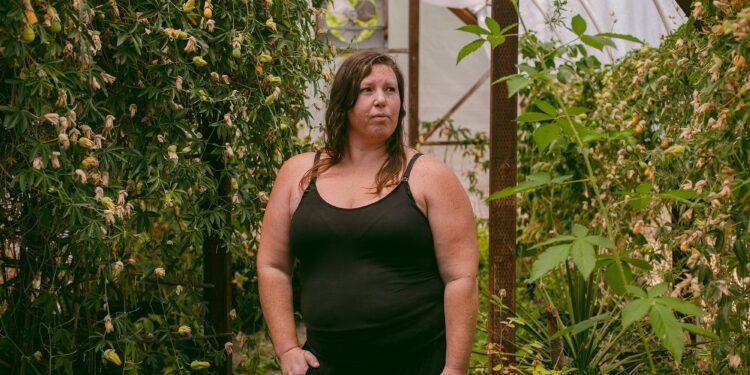Susan Horton, a White woman wearing a black tank top and black pants, poses for a portrait inside a greenhouse.