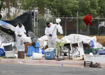 Two city workers, wearing black caps, black gloves and white coveralls, hold plastic bags as they “sweep” the remains of a homeless encampment.