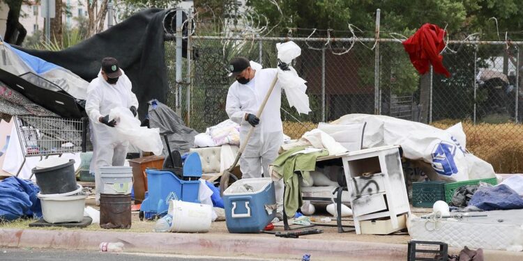 Two city workers, wearing black caps, black gloves and white coveralls, hold plastic bags as they “sweep” the remains of a homeless encampment.