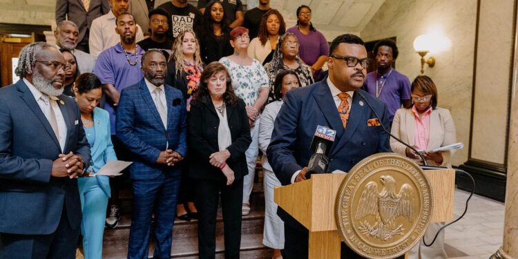 A photo of Kabir Karriem, a Black man, speaking at a podium while wearing a blue suit and glasses. Karriem has short black hair and a mustache. More than 20 people stand behind him.
