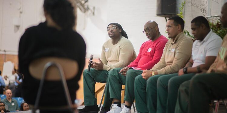 A Black man wearing glasses, dark green prison pants and a tan sweater is holding a microphone as he asks a question to a woman in the foreground who is sitting on a chair and her back turned to the camera. Alongside the man sit four other men in the same green prison pants and a variety of shirts. Members of the audience look on from their blue-colored seats.