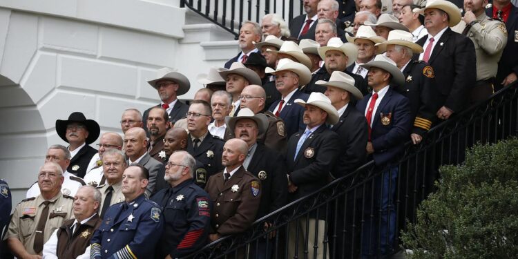 Dozens of sheriffs in uniform, some wearing cowboy hats, some wearing suits, stand on the steps of an angled staircase outside of The White House.