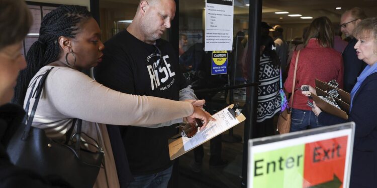 A White man wearing a black T-shirt, a gray long-sleeved shirt and blue jeans, holds a clipboard with a voter registration form. A Black woman wearing a light gray cardigan and holding a black purse, points at the registration form as they stand at the entrance of a building. People are standing in front of them in a line.