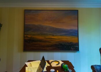 A painting showing a mountainside, orange clouds and blue sky hangs on a wall as light comes in from the right side. A table with a laptop and some papers is visible at the bottom of the photo.