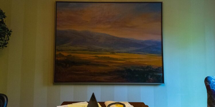 A painting showing a mountainside, orange clouds and blue sky hangs on a wall as light comes in from the right side. A table with a laptop and some papers is visible at the bottom of the photo.