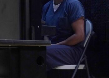 A man wearing a blue uniform sits on a metal folding chair in a small room.