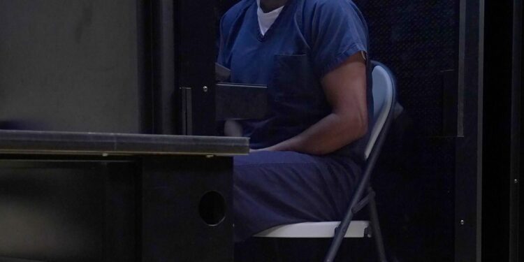 A man wearing a blue uniform sits on a metal folding chair in a small room.