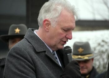A White man with white hair speaks into a microphone outdoors, with two White men in uniform hats visible behind him.