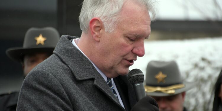 A White man with white hair speaks into a microphone outdoors, with two White men in uniform hats visible behind him.