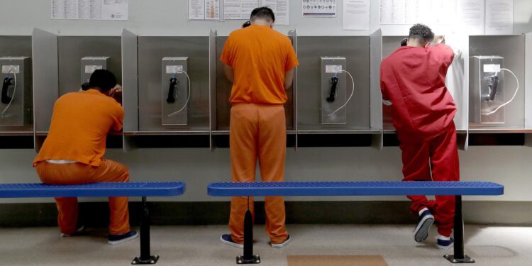 Three men, wearing orange and red uniforms, talk on telephones in a payphone booth area.
