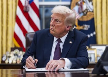 Trump, a White man in a navy suit and a red and navy tie, signs a document while sitting in the Oval Office.