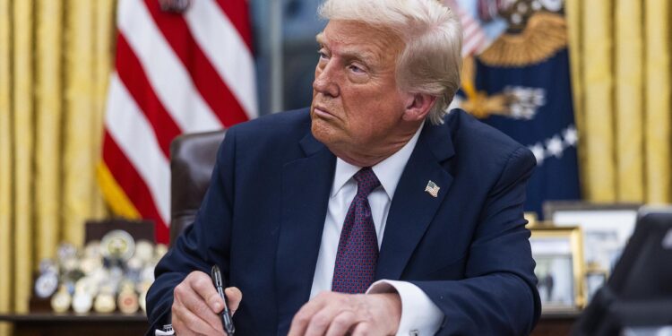 Trump, a White man in a navy suit and a red and navy tie, signs a document while sitting in the Oval Office.