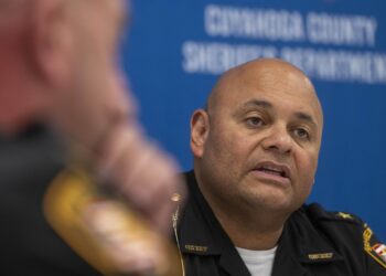 A man with medium-toned skin, wearing a uniform, sits in front of a blue wall with the words “Cuyahoga County Sheriff’s Department” on it. 
