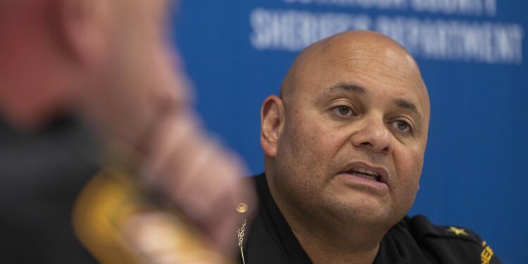 A man with medium-toned skin, wearing a uniform, sits in front of a blue wall with the words “Cuyahoga County Sheriff’s Department” on it. 
