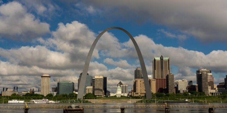 The Mississippi River is visible in the foreground of a photo, with the Gateway Arch in the center.  Buildings are in the background, and a blue sky with clouds is visible.