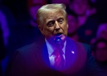A photograph of President Trump, a White man in a suit and tie, speaking at a podium behind pink and blue orbs of light.