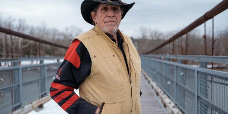 George Hall, an older White man wearing a hat with a brim and a tan vest and long sleeved jacket, stands on a bridge.