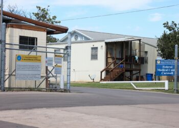 Metal fencing can be seen in front of two structures, one on the right with a short wooden staircase, and one on the left with beige siding. A sign in front of the building on the right reads: Federal Medical Center Carswell.