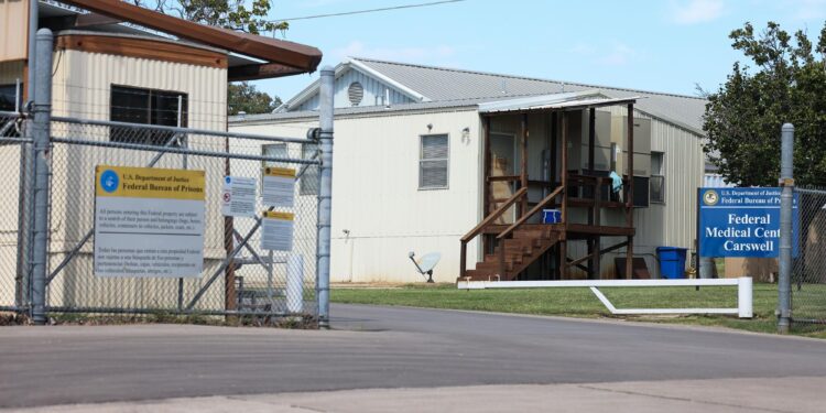 Metal fencing can be seen in front of two structures, one on the right with a short wooden staircase, and one on the left with beige siding. A sign in front of the building on the right reads: Federal Medical Center Carswell.