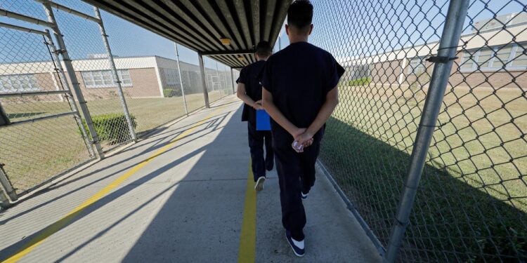 Two men wearing black uniforms clasp their hands behind they walk through a fenced pathway.