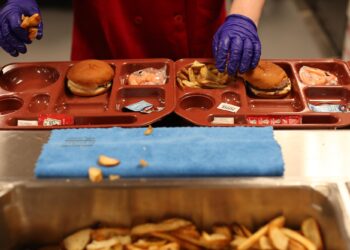 An incarcerated person wearing food preparation gloves adds French fries to a food tray that contains a hamburger and a small bag of baby carrots.