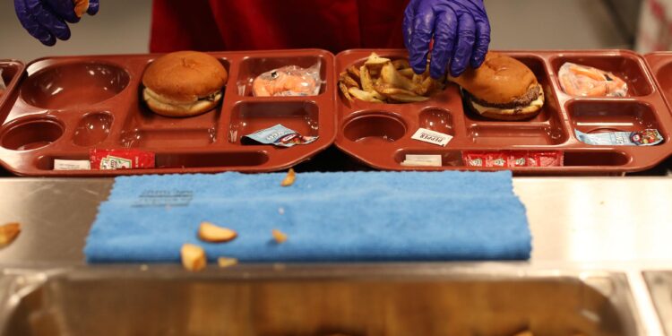 An incarcerated person wearing food preparation gloves adds French fries to a food tray that contains a hamburger and a small bag of baby carrots.