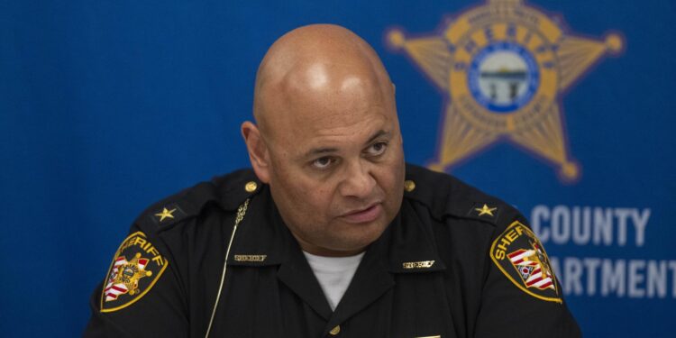 Sheriff Harold Pretel, a man with medium-toned skin, wearing a sheriff's uniform, sits in front of a blue background with a sheriff's star printed on it.