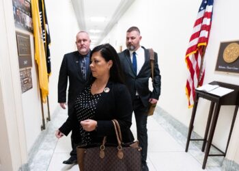 A woman carrying a brown handbag walks in the white hallways of the Rayburn House Office Building with two men standing behind her.