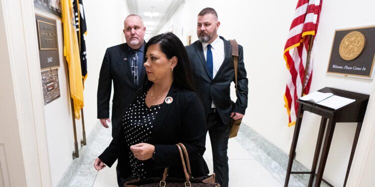 A woman carrying a brown handbag walks in the white hallways of the Rayburn House Office Building with two men standing behind her.